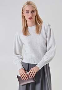 Donna bionda con un maglione bianco a trama e una gonna plissettata grigia, che tiene una pochette metallica contro uno sfondo semplice.
