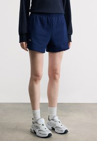 Άτομο που φοράει σορτς σε σκούρο μπλε χρώμα, λευκές αθλητικές κάλτσες και λευκά αθλητικά παπούτσια, στέκεται σε γκρι πάτωμα δίπλα σε λευκό τοίχο.