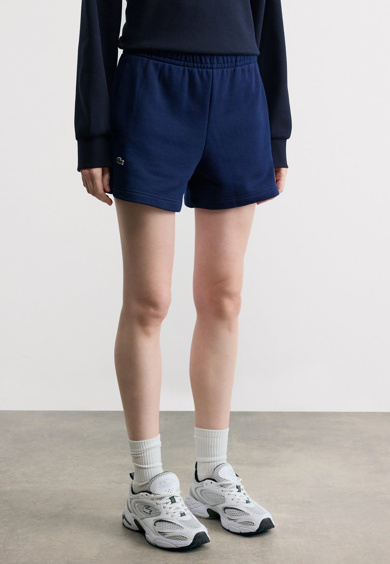 Άτομο που φοράει σορτς σε σκούρο μπλε χρώμα, λευκές αθλητικές κάλτσες και λευκά αθλητικά παπούτσια, στέκεται σε γκρι πάτωμα δίπλα σε λευκό τοίχο.