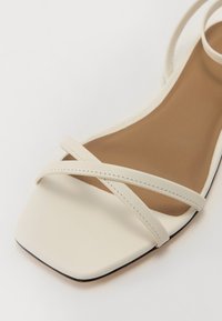 White faux leather sandal with square toe, featuring crisscross straps and a smooth brown insole, set against a light background.