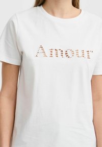 Bílý bavlněný tričko s krátkými rukávy, vyznačující se laserově řezaným textem "Amour" ve pruhovaném vzoru. Hladká textura a klasický kulatý výstřih.
