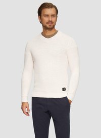 Mann in einem weißen, langärmeligen V-Ausschnittpullover mit einem kleinen schwarzen Etikett, dunkler Hose, der mit einer Hand in der Tasche vor einem neutralen Hintergrund steht.