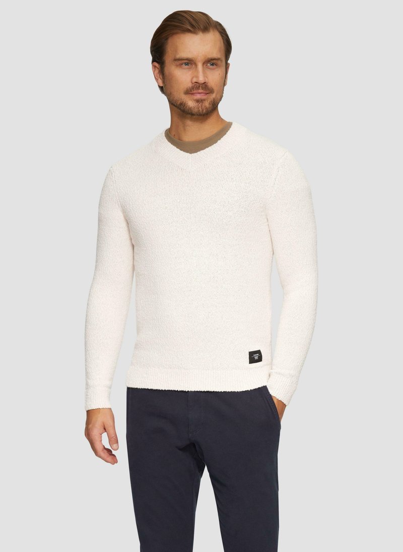 Mann in einem weißen, langärmeligen V-Ausschnittpullover mit einem kleinen schwarzen Etikett, dunkler Hose, der mit einer Hand in der Tasche vor einem neutralen Hintergrund steht.
