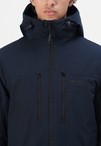 Mann iført en marineblå hettejakke med to vertikale brystglidelåser og "DENIM TOM TAILOR" merkevare på høyre bryst.