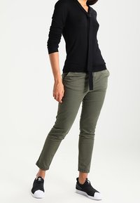 Czarny top z długim rękawem z detalem w postaci wiązania, oliwkowe slim fit spodnie oraz czarne trampki z białymi podeszwami. Gładka faktura, casualowy design.