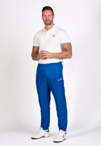 Weißes Polohemd, blaue Sporthose mit elastischem Bund und weißen Sneakers. Die Hose hat ein Logo auf der linken Seite. Lässige Passform.