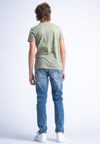 Lichtgroene katoenen T-shirt met een ronde "Petrol Industries" print in heldergeel op de achterkant, gecombineerd met lichtblauwe denim jeans en witte sneakers.