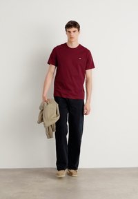 Mørkerød T-shirt med et lille logo, parret med mørke jeans. Modellen holder en lys beige jakke og har lyse sko på. Ensfarvet baggrund.