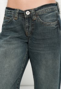 Calças de ganga em tom azul escuro com uma textura ligeiramente desbotada, apresentando um fecho de botão padrão e detalhes com costuras nos bolsos.