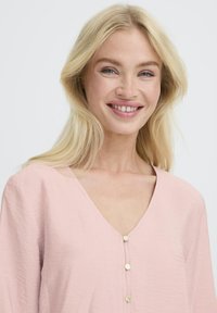 Naeratav naine pikade blondide juustega, seljas heledat roosa V-kaelusega pluus, millel on kolm väikest kuldset nuppu, ühtlasel taustal.