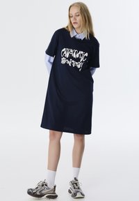 Marine-blauwe T-shirt jurk met een losse pasvorm, voorzien van witte abstracte tekst op de voorkant. Inclusief een gelaagd blauw en wit gestreept shirt eronder.