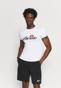 Bijela kratkouhana majica s crnim "ellesse" logotipom i šarenim dizajnom luka. Uparena s crnim šortsevima s bijelim Nike logotipom.
