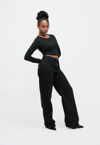 Svart kort topp med lange ermer kombinert med høyhøyde svarte vide bukser. Modellen har på seg svarte spisse hæler; satt mot en enkel hvit bakgrunn.