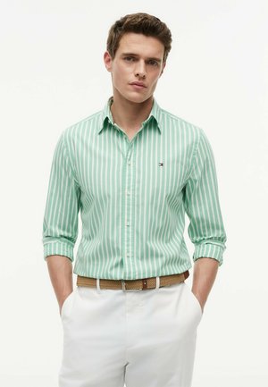 Junger Mann mit kurzen braunen Haaren, der ein hellgrün-weiß gestreiftes Langarmshirt, weiße Hosen und einen gewebten braunen Gürtel trägt.