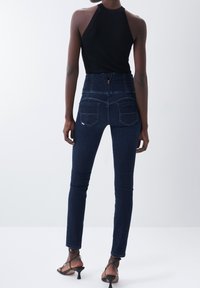 Jeans de tiro alto azul oscuro con una textura suave, diseño ajustado y dos bolsillos traseros. Combinados con un top halter negro ajustado.