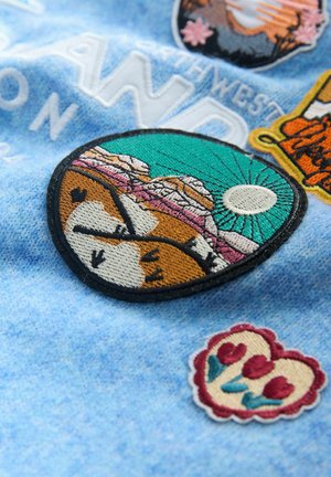 Embroidered patches on a blue fabric background; one features mountains and sun in green, brown, and pink, while another shows flowers in a heart shape.