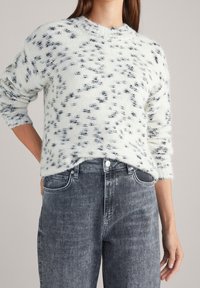 Maglione bianco lavorato a maglia con macchie nere, colletto rotondo e maniche lunghe, abbinato a jeans in denim grigio a vita alta. Materiale morbido e testurizzato.