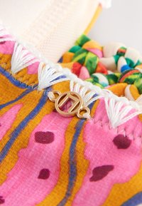 Gros plan sur un tissu coloré aux motifs roses et moutarde, avec une couture en frange blanche, un pendentif circulaire doré, et un textile tressé multicolore en arrière-plan.
