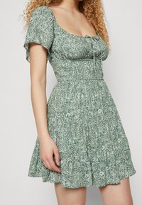 Žena s kudrnatými vlasy předvádí zelené květinové šaty s krátkým rukávem a p řiléhavým pasem, stojící na neutrálním pozadí.