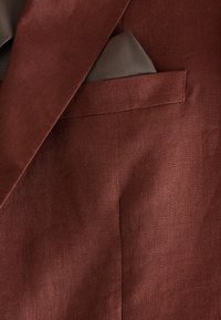 Gros plan sur le revers d'un blazer marron avec une petite poche poitrine tenant un mouchoir gris, montrant un tissu texturé et des détails de couture.