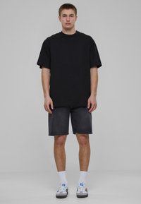 Zwarte oversized t-shirt met korte mouwen, gecombineerd met donkere denim shorts. Model draagt witte sokken en grijze sneakers met zwarte accenten.