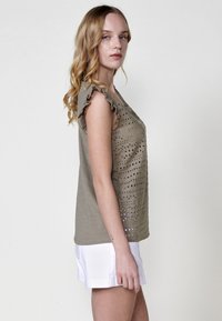 Top sin mangas color caqui con diseño de ojales, con volantes en los hombros y textura suave, combinado con pantalones cortos blancos, vista lateral.