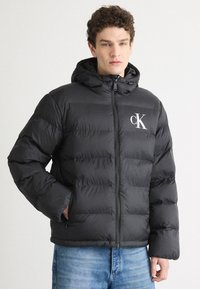 Schwarze wattierte Jacke mit durchgehendem Reißverschluss, Kapuze und weißem Logo. Sie hat ein gestepptes Design und einen lockeren Schnitt.