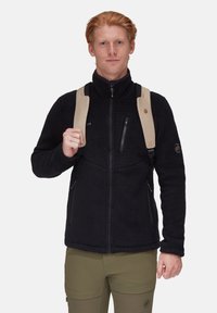 Schwarze Fleecejacke mit hohem Kragen, Reißverschluss-Brusttasche und Seitentaschen. Beige Riemen und ein Logo-Patch betonen das Design.