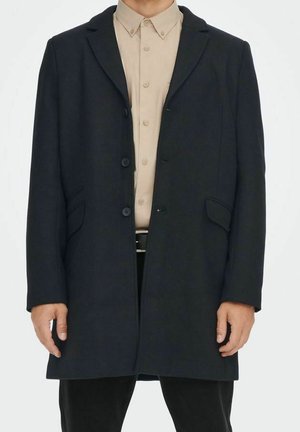 Homme portant un manteau noir à simple boutonnage sur une chemise beige à boutons et un pantalon noir, debout devant un fond blanc.