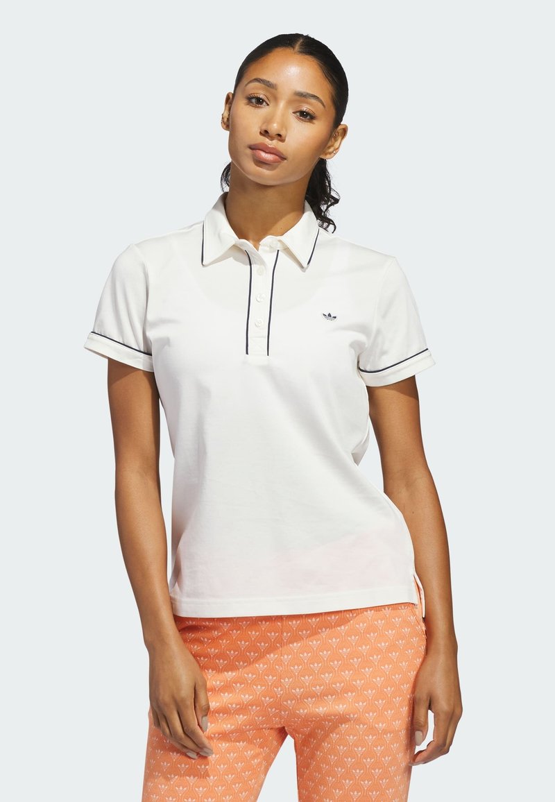 Femme aux cheveux foncés portant un polo blanc à manches courtes avec bordure noire et un pantalon à motifs orange, debout devant un fond clair uni.