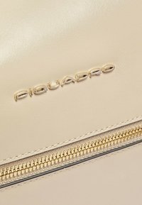 Beige leren tas met een rits in goudkleur. Voorzien van een geperst "Piquadro"-logo en een gladde textuur, met fijne stiksels langs de rand.