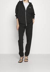 Musta vetoketjullinen huppari, jossa on etutasku, yhdistettynä mustiin verkkareihin. Molemmissa on sileä pinta, ja ne sopivat täydellisesti mustien remmisaappaiden kanssa.