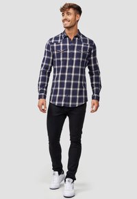 Navy blue flannel shirt with white plaid pattern, featuring a button-down collar, two chest pockets, and long sleeves. Black fitted jeans and white sneakers.