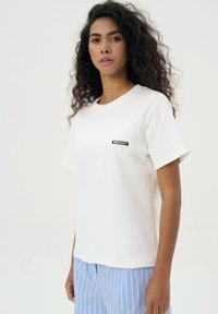 Biała bawełniana koszulka z okrągłym dekoltem i krótkimi rękawami, z małą czarną łatą logo po lewej stronie.