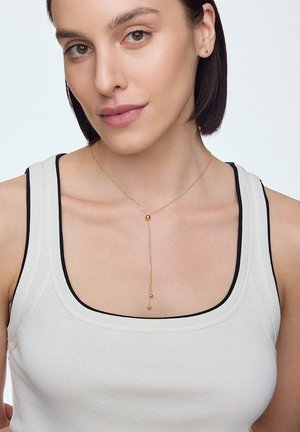 Złoty naszyjnik z delikatnym łańcuszkiem, z centralną perłą i dwiema dolnymi perłami, noszony na jasnym topie z czarnym wykończeniem.