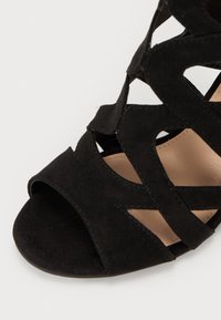 Zwarte suède hoge hak schoenen met uitgesneden ontwerp, open teen en een gladde beige binnenzool. Getextureerde buitenkant met accentbandjes.
