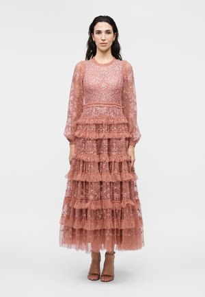 Kobieta o długich ciemnych włosach nosi różową koronkową sukienkę midi z falbankami i przezroczystymi bufkami na rękawach, stojącą na jednolitym tle.