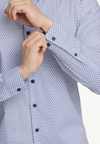 Camicia a fantasia blu chiaro con un design geometrico, caratterizzata da bottoni blu e polsini abbottonati. Il tessuto appare liscio con una leggera lucentezza.