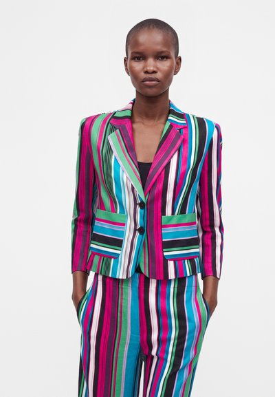 Femme portant un blazer rayé coloré et un pantalon assorti avec des rayures verticales et diagonales vertes, roses, bleues, noires et blanches.