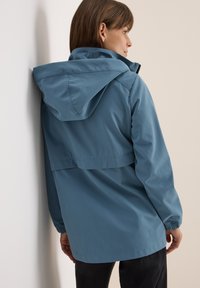 Veste imperméable bleu clair avec capuche, coutures visibles et poignets élastiques. Présente une coupe décontractée et une longueur mi-longue.