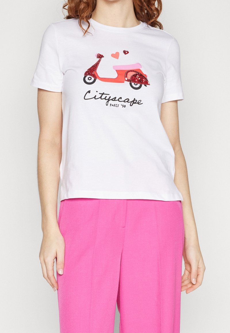 Femme portant un t-shirt blanc avec un scooter rouge et un graphique de cœurs et le texte "Cityscape Paris '90", associé à un pantalon rose vif.