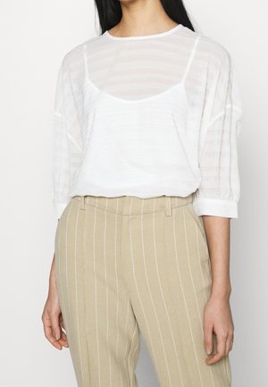 Femme portant un chemisier blanc transparent rayé sur un caraco blanc et un pantalon beige taille haute à fines rayures blanches verticales.