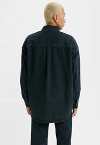 Camisa de mezclilla oscura con un corte relajado, costuras visibles, puños con botones y dobladillo curvado, vista desde la espalda.