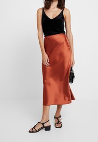 Top in velluto nero con spalline a spaghetti, gonna midi in raso color ruggine, sandali con tacco a blocco neri, piccola borsa nera, texture morbide.