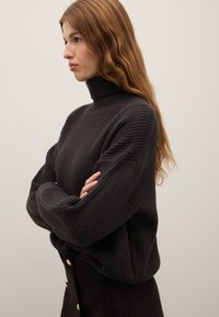 Dunkelgrauer Rollkragenpullover mit gerippter Textur, lockerer Passform und überschnittenen Schultern. Kombiniert mit einem gemusterten, dunklen Rock mit goldenen Knöpfen.