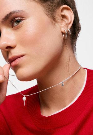 Joven mujer con suéter rojo sosteniendo un collar plateado con un colgante de perla y llave cerca de su rostro, llevando pendientes de aro plateados.