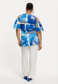 Bluse mit floralem Muster in Blau und Weiß mit kalten Schultern, kombiniert mit weißen weit geschnittenen Hosen und blauen Blockabsätzen.