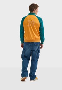 Färgglad bomberjacka med tealärmar och en orange kropp, med en stor "39" tryckt på baksidan. Matchas med lösa denim cargo-byxor.