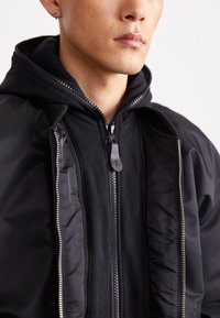 Black bomber jacket with a zip closure, featuring a hood, contrasting inner lining, and exposed zippers. Smooth fabric texture.