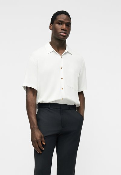 Homme portant une chemise blanche à manches courtes boutonnée, rentrée dans un pantalon noir, avec une main dans la poche, devant un fond uni.